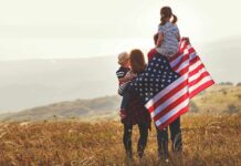 Family in field with American flag, children on shoulders.