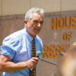 Man speaking with microphone outside building with lettering