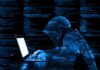 Hooded figure using laptop with code background.