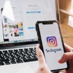 Person holding a smartphone displaying the Instagram logo with a laptop in the background