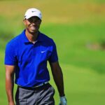 Tiger Teases Masters Shocking Comeback A smiling golfer wearing a blue shirt and white cap on a green golf course