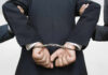Person handcuffed in suit, escorted by two others.