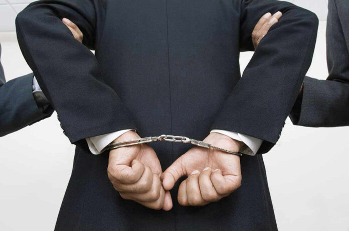 Person handcuffed in suit, escorted by two others.