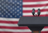 Podium with microphones in front of American flag.