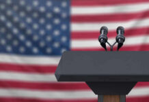 Podium with microphones in front of American flag.