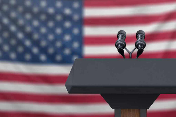 Podium with microphones in front of American flag.