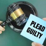 A wooden gavel next to a hand holding a blue card that says 'PLEAD GUILTY'
