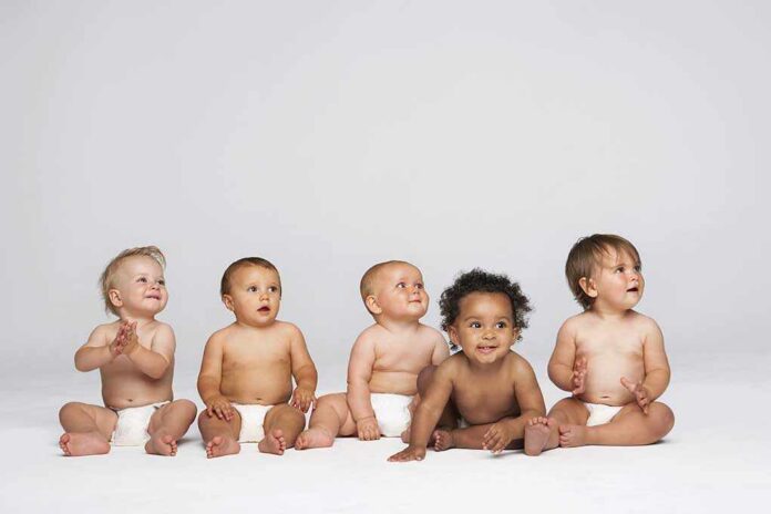 Row,Of,Multiethnic,Babies,Sitting,Side,By,Side,Looking,Away Five diverse babies sitting on a neutral background, displaying playful expressions