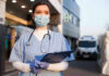 Fake Nurses Still Treating Patients Nurse in scrubs and mask outside hospital holding clipboard.