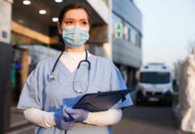 Fake Nurses Still Treating Patients Nurse in scrubs and mask outside hospital holding clipboard.