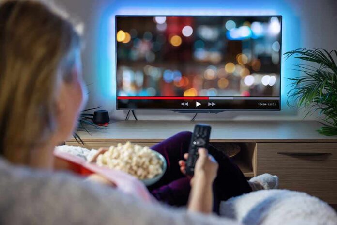 shutterstock_1966720738 Person sitting on a couch watching a movie on a TV with popcorn in hand