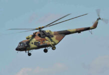 Army Black Hawks Swarm Kid-Filled Camp Camouflage military helicopter in flight against blue sky.