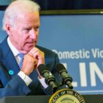 man at podium speaking with hands clasped near chin