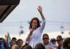 Woman speaking at outdoor event with raised hand.