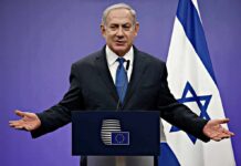 man in suit speaking at a podium with Israeli flag