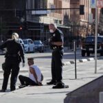 ICE-Linked Church Protest: Legal Chaos Unleashed Police officers detain a seated man on a city sidewalk