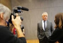 Kennedy Torches Dems, GOP Crowd ERUPTS Man in a gray suit speaks to a reporter during an interview