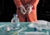 Handcuffed person in orange jumpsuit behind medical vials and IV equipment
